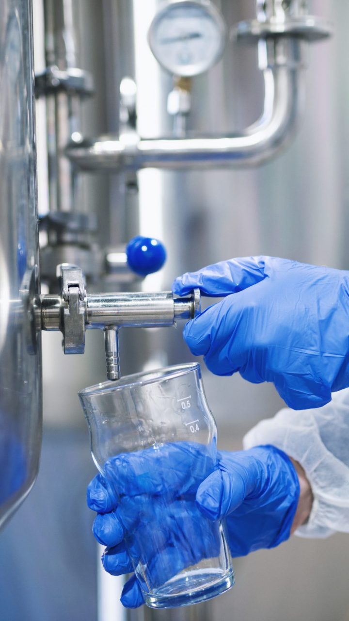 Close up view of hands in protective gloves opening tap valve and filling glass with samples from industrial machine reservoir.