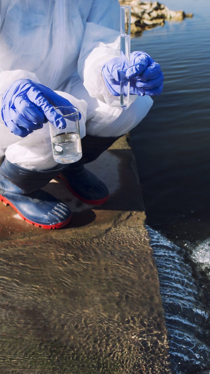 Unrecognizable ecologist standing where sewage waste water meets the river and taking samples to determine level of contamination and pollution.